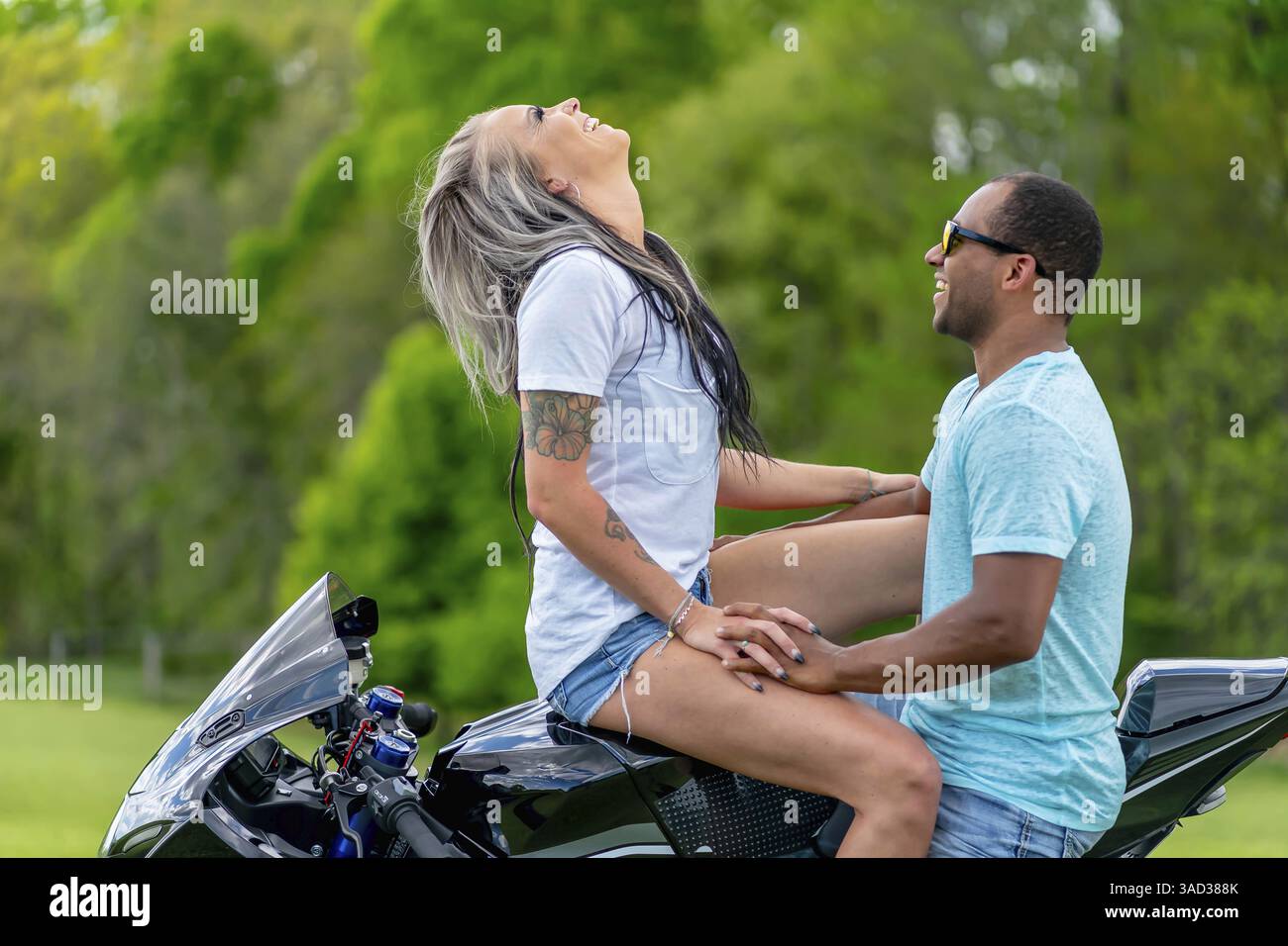 Una coppia multi-razziale posa amorevolmente accanto alla propria moto in un campo lussureggiante e verde. Abbracciati, sorridono con orgoglio, catturando entrambi i loro legami profondi Foto Stock