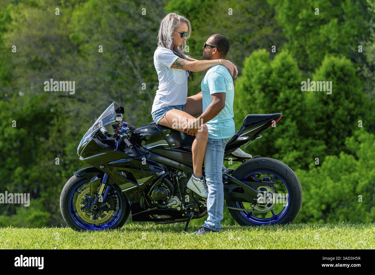 Una coppia multi-razziale posa amorevolmente accanto alla propria moto in un campo lussureggiante e verde. Abbracciati, sorridono con orgoglio, catturando entrambi i loro legami profondi Foto Stock