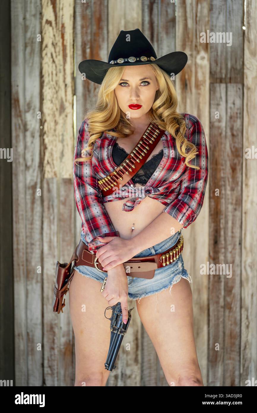 Una bella e sicura donna bionda in un classico abito da cowboy si erge a tutta altezza, in posa con una scintillante pistola sull'anca e un fucile appoggiato sulla sua shou Foto Stock