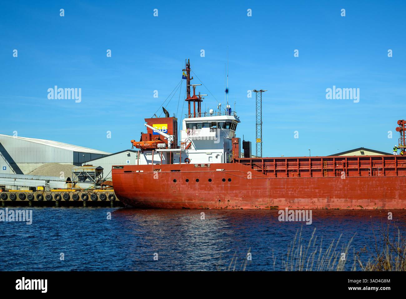 Una nave da carico rossa è ancorata in un porto affollato sotto un cielo azzurro. I magazzini e le gru circostanti indicano che sono in corso operazioni di spedizione attive Foto Stock