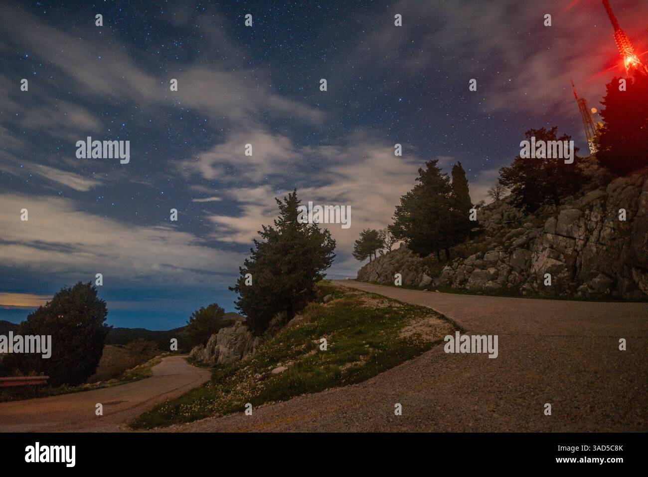 Una strada tortuosa su una collina rocciosa sotto un cielo stellato, con alberi e luci lontane visibili. Foto Stock
