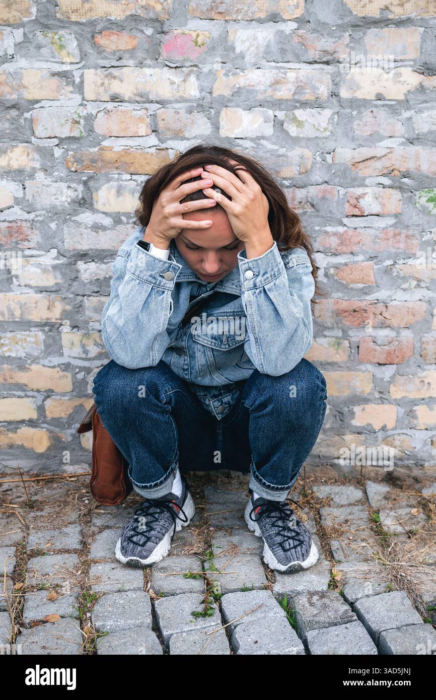 Donna con dolore emotivo seduta e con la testa in mano contro il muro di mattoni per strada Foto Stock