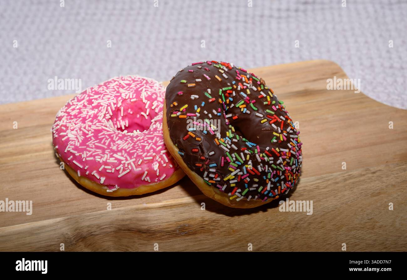 Due deliziose ciambelle si trovano su una tavola di legno, una ricoperta di glassa rosa e spruzzi bianchi, e l'altra con una ricca ricopertura di cioccolato e una vivace m Foto Stock
