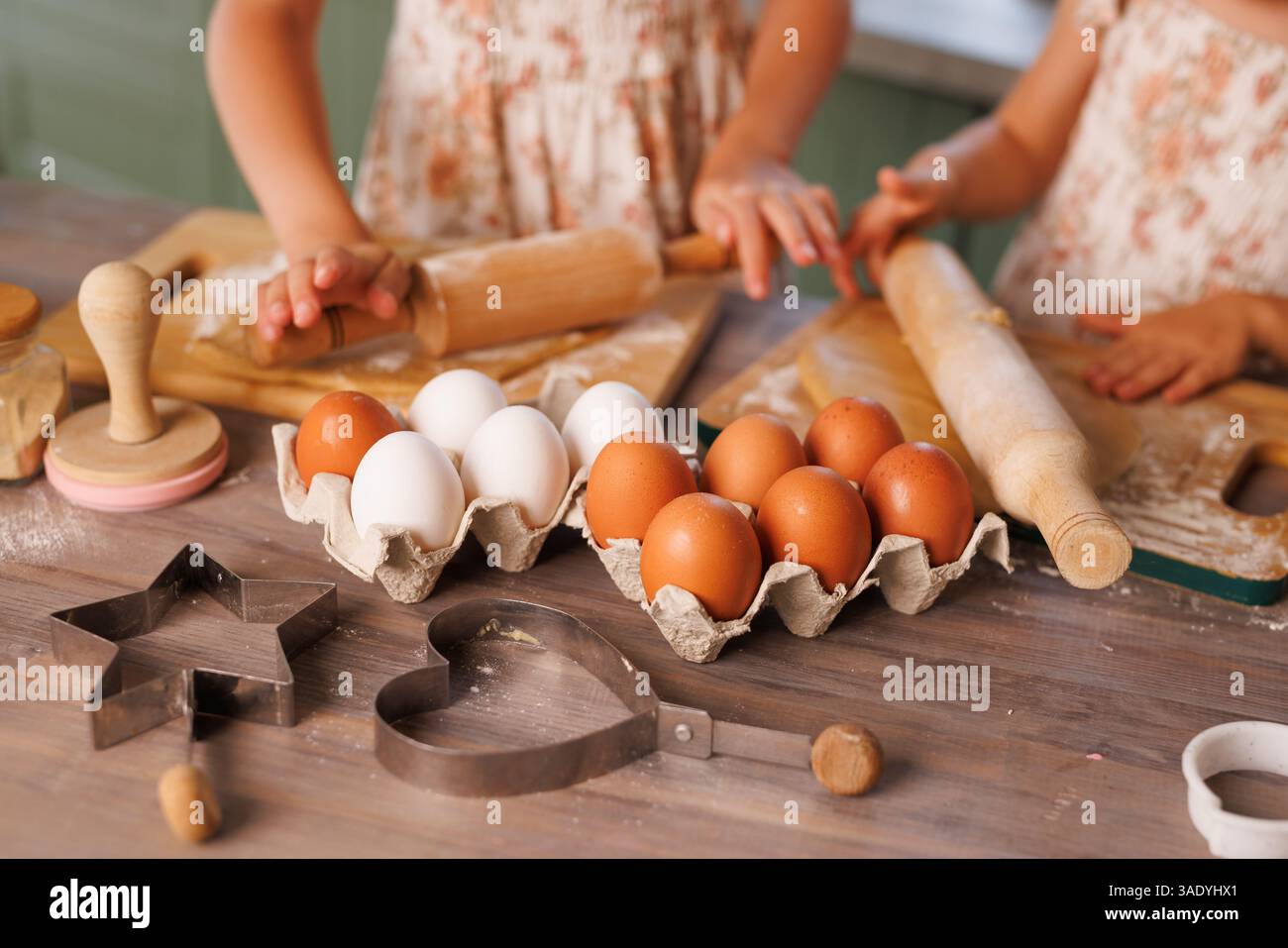 I bambini cuocono insieme i biscotti, arrotolano l'impasto e usano le taglierine con uova fresche e farina su un tavolo rustico. Foto Stock