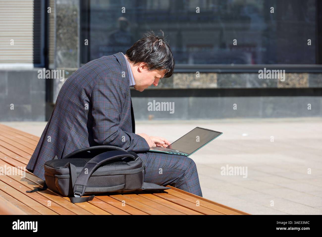 Uomo con un costume da lavoro che lavora con un computer portatile su una panchina in città Foto Stock