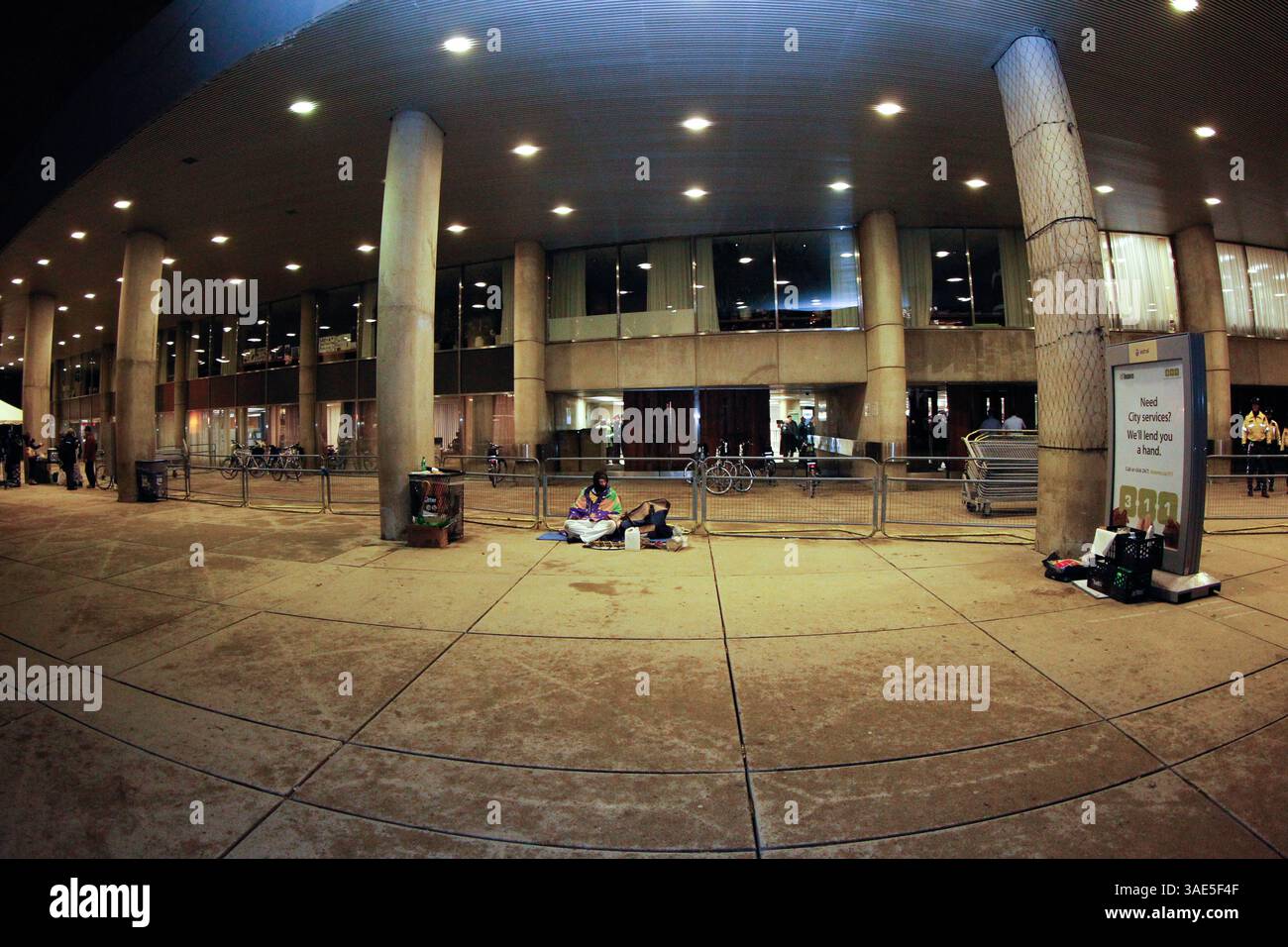 17 gennaio 2012 - Toronto, Ontario, Canada - Un protestatore solitario siede accanto a un annuncio pubblicitario "Need City Services - We'll Lend You A Hand"...mentre i consiglieri della città di Toronto hanno discusso del budget 2012 della città, la polizia di Toronto è stata costretta a mettere il Municipio in blocco per far fronte a una protesta sempre più violenta contro i tagli in Nathan Phillips Square. ..Un aiuto della polizia in tutta la città è stato rilasciato quando è diventato chiaro che la manifestazione pacifica con diverse centinaia di manifestanti stava diventando acida. Quattro manifestanti maschili sono stati presi in custodia, accusati di essere assemblati illegalmente e di ostacolare un ufficiale di polizia. ..ma Foto Stock