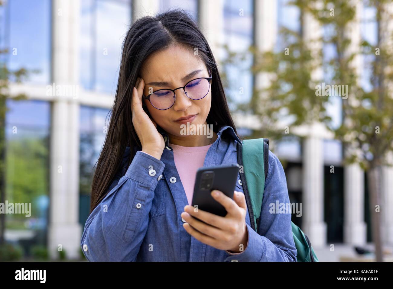 Una donna asiatica che indossa gli occhiali sta guardando il suo telefono con un'espressione preoccupata. Sembra stressata, forse a causa di un messaggio. Foto Stock