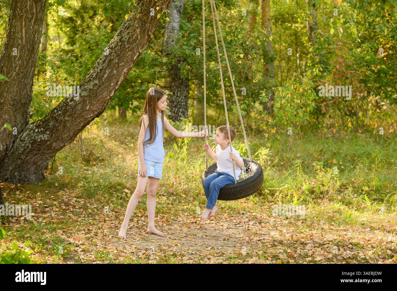 La sorella dondola il ragazzo seduto comodamente su una ruota, giocando e chiacchierando insieme. Bambini che oscillano su pneumatici per auto in gomma. Vacanze in campagna, Sum Foto Stock