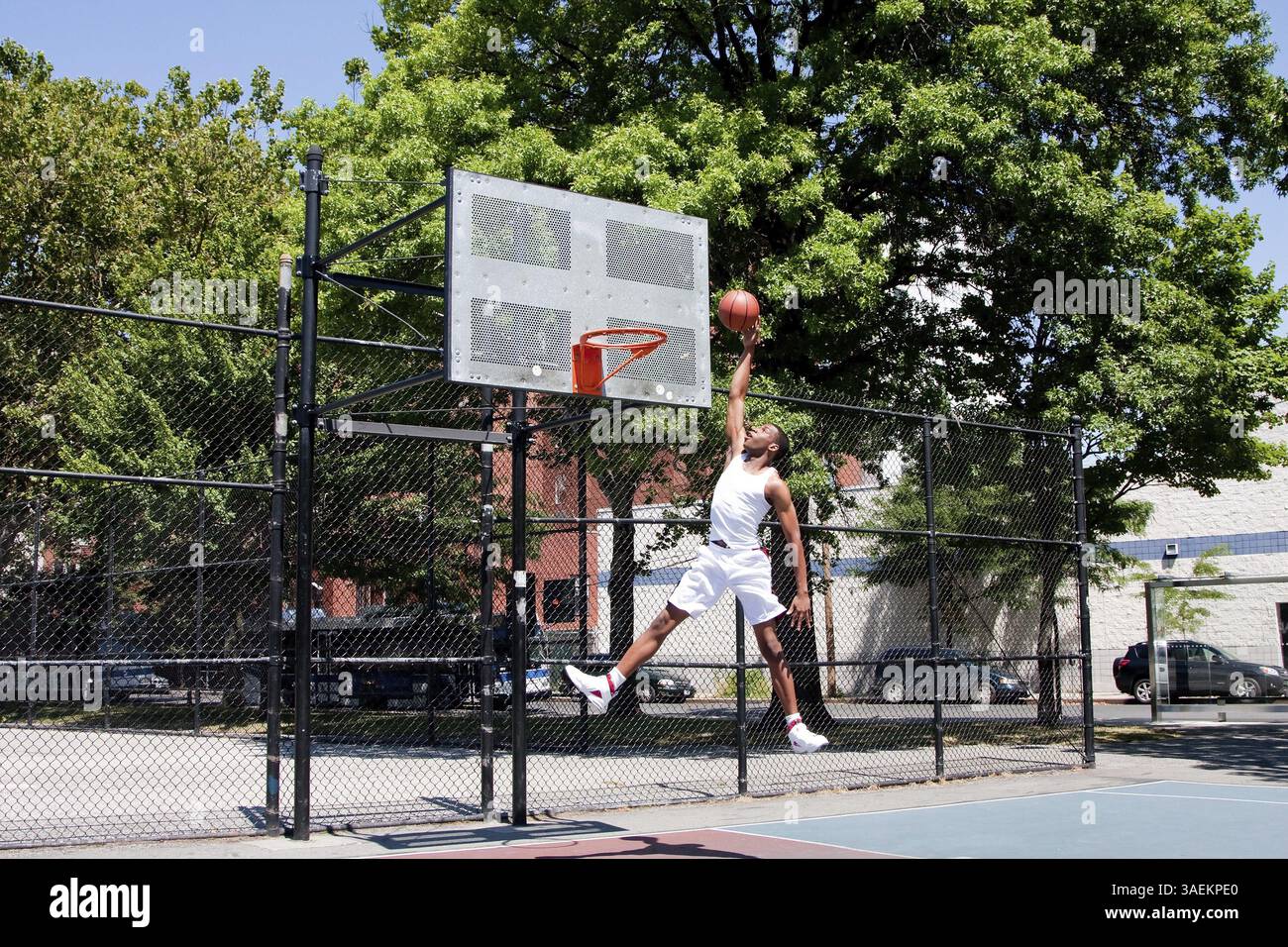 Sporty bello afro-americano di uomo vestito di bianco di saltare in aria per raggiungere il cesto mentre si gioca a basket in un tribunale aperto su una somma Foto Stock