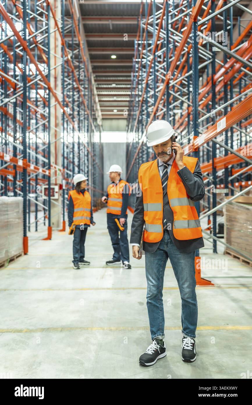 Il responsabile del magazzino indossa il giubbotto di sicurezza e il casco utilizzando il telefono cellulare, coordinando le operazioni logistiche con il suo team in un grande centro di distribuzione Foto Stock