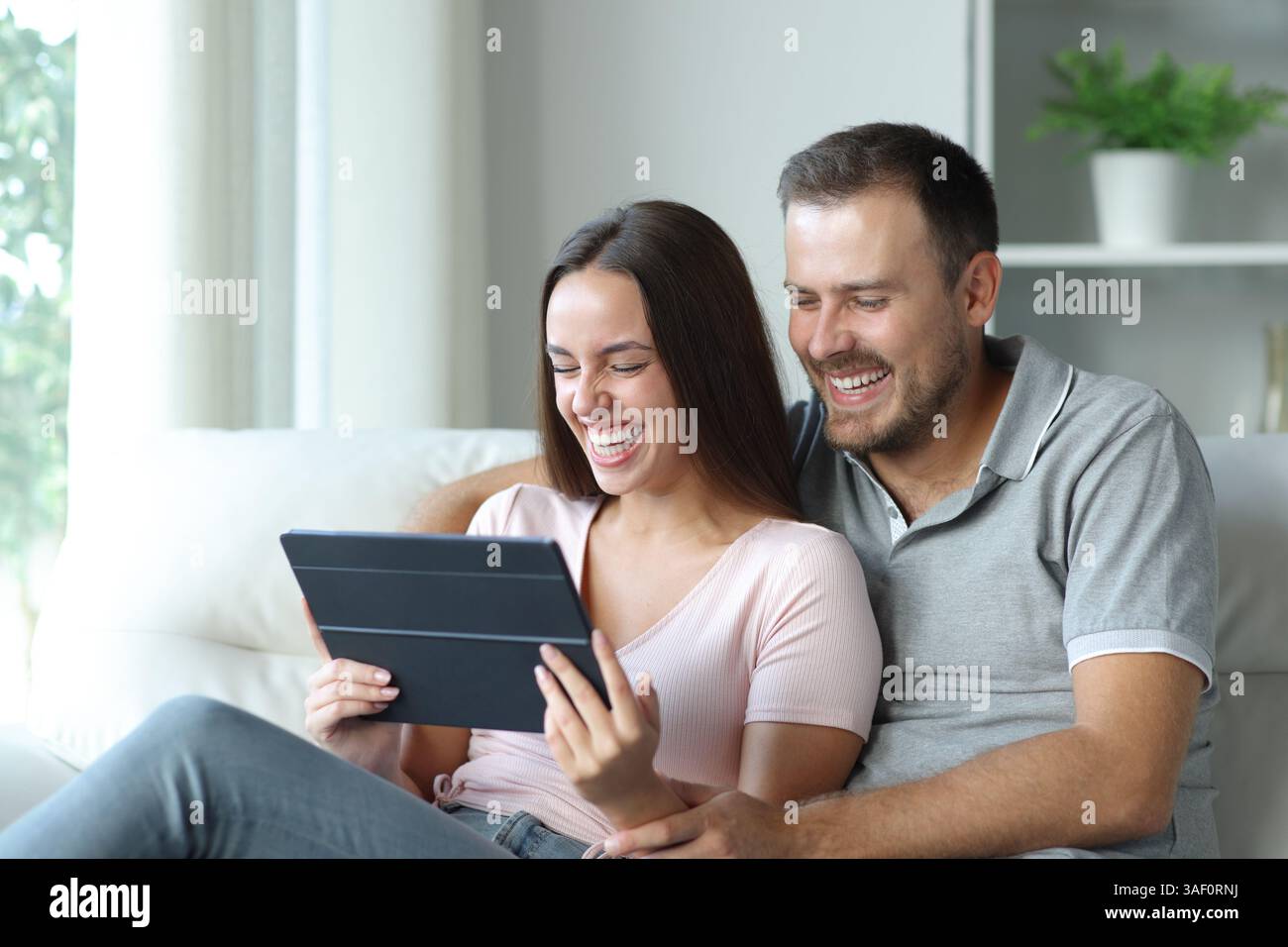 Coppia felice che controlla contenuti divertenti sul tablet seduto su un divano a casa Foto Stock