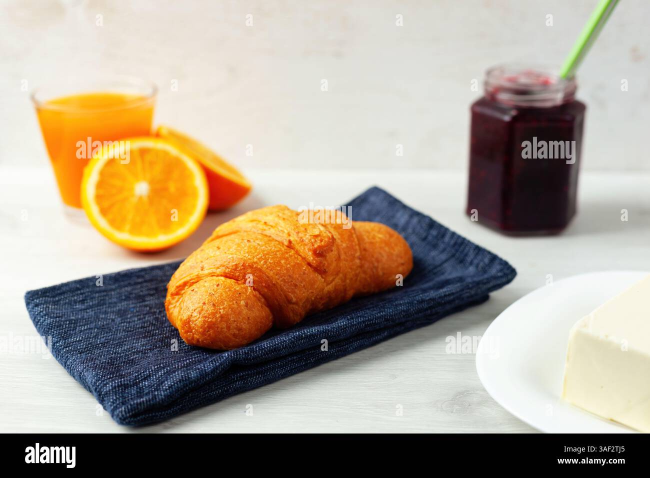 Croissant appena sfornato appoggiato su un tovagliolo blu, accompagnato da succo d'arancia rinfrescante, marmellata dolce e burro cremoso, per creare una deliziosa e nutriente colazione Foto Stock