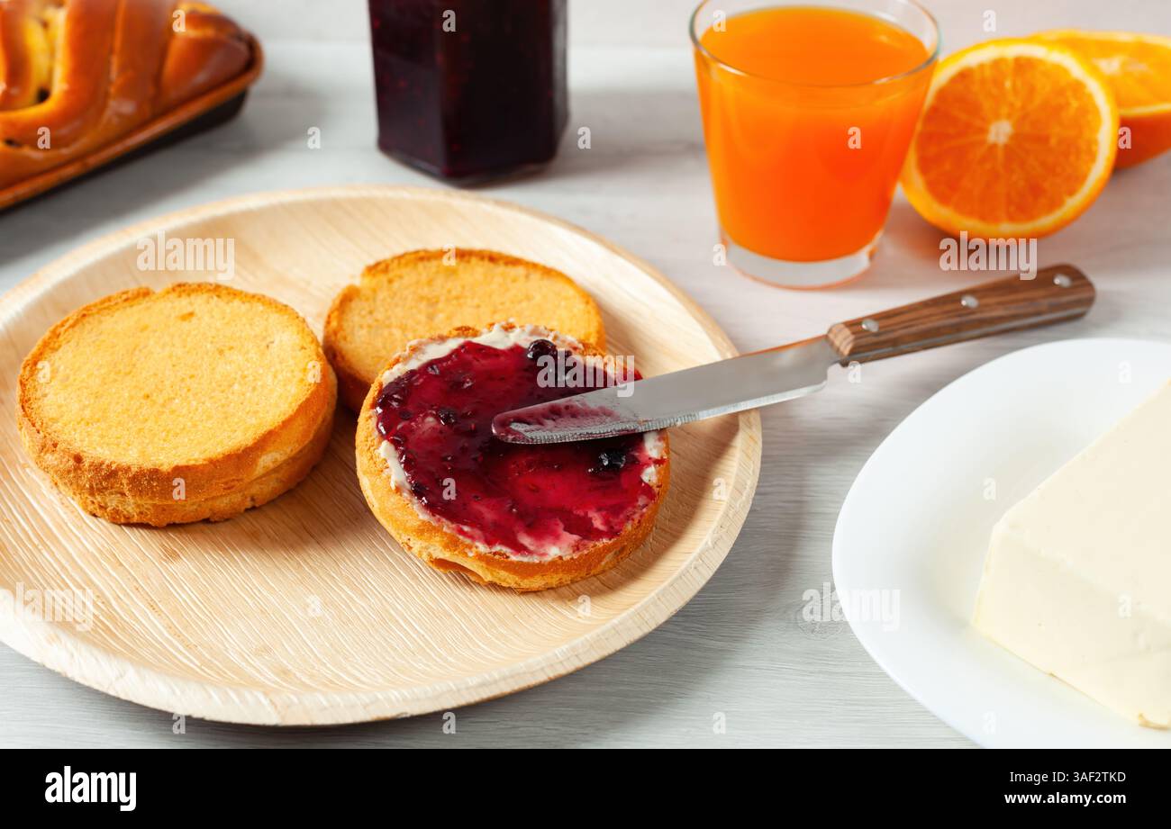 Spalmare la marmellata di mirtilli sul pane tostato con un coltello, accompagnato da succo d'arancia, burro e pane intrecciato, crea una scena di colazione sana e deliziosa Foto Stock
