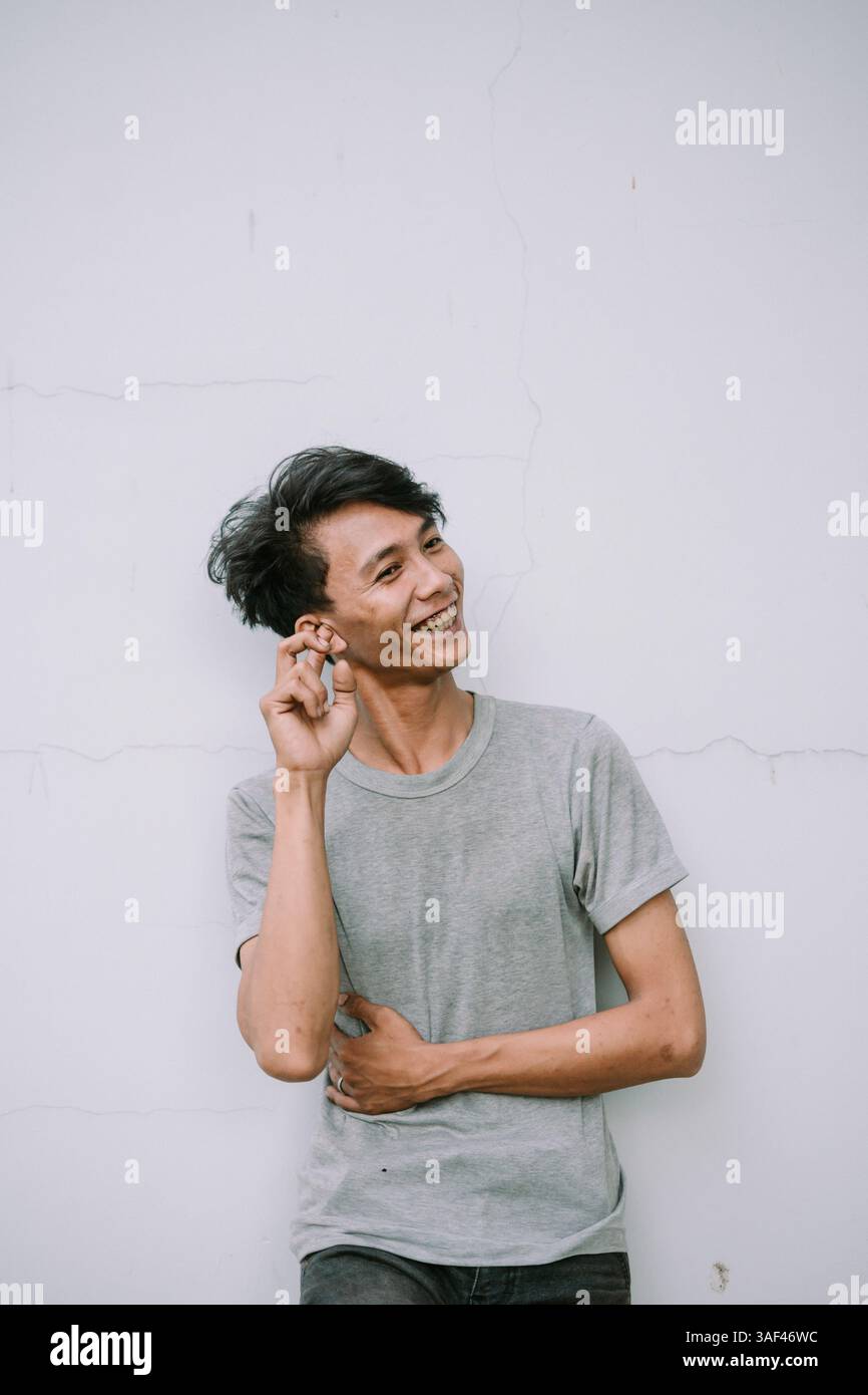 Giovane sorridente che posa accanto al muro con camicia grigia ed espressione felice Foto Stock