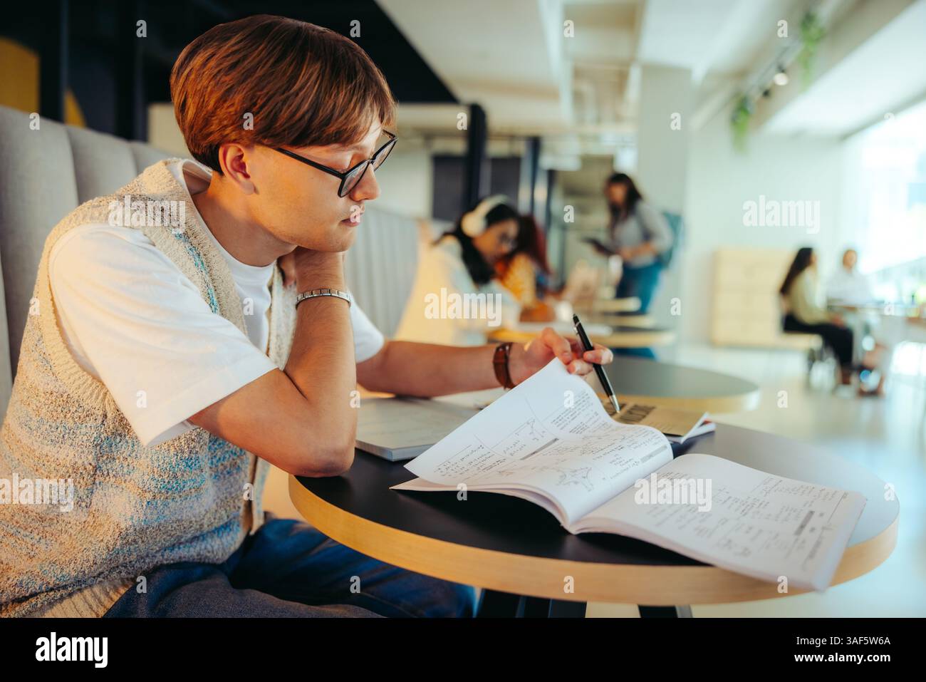 Uno studente di scuola superiore maschile sta studiando intensamente in una biblioteca, circondato da colleghi in un ambiente di apprendimento collaborativo. Foto Stock