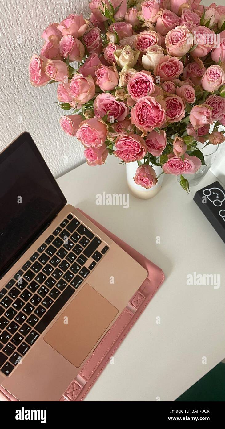 Un computer portatile in oro rosa e un mazzo di rose rosa su una scrivania bianca. Spazio di lavoro femminile, luce soffusa ed estetica delicata per la creatività Foto Stock