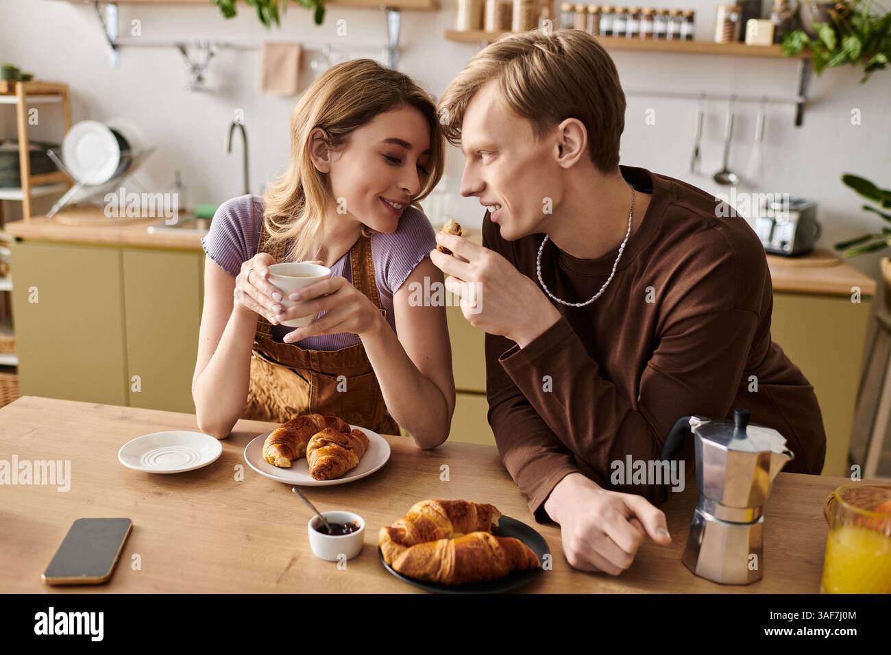 Una giovane coppia condivide con gioia la colazione con croissant e bevande calde nella loro luminosa cucina. Foto Stock