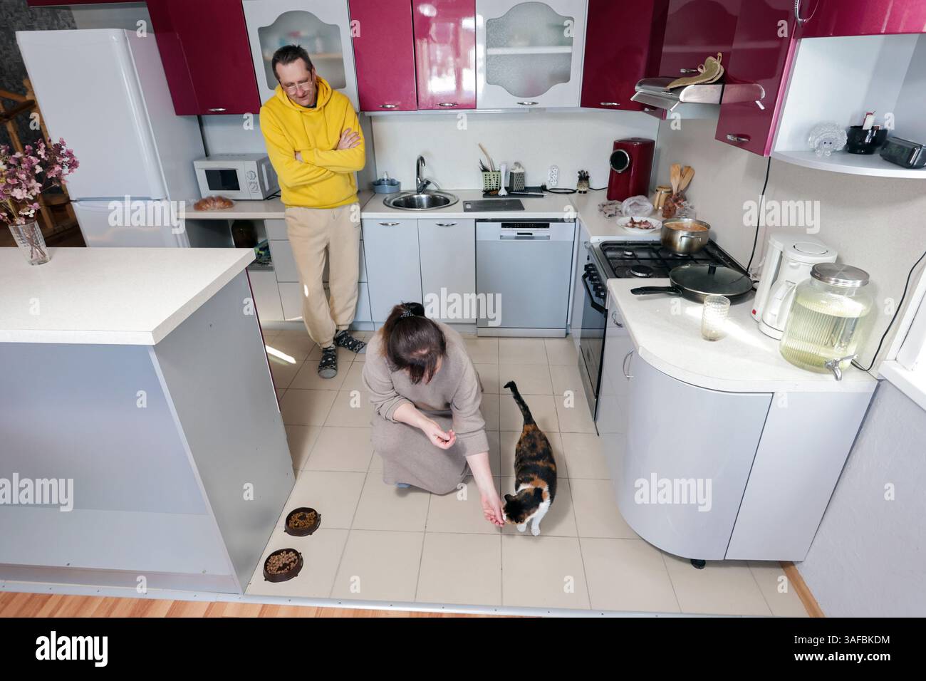 L'uomo osserva la donna mentre dà da mangiare al gatto sul pavimento della cucina piastrellata, condividendo un'interazione tranquilla piena di cura e calma in un ambiente familiare. Foto Stock