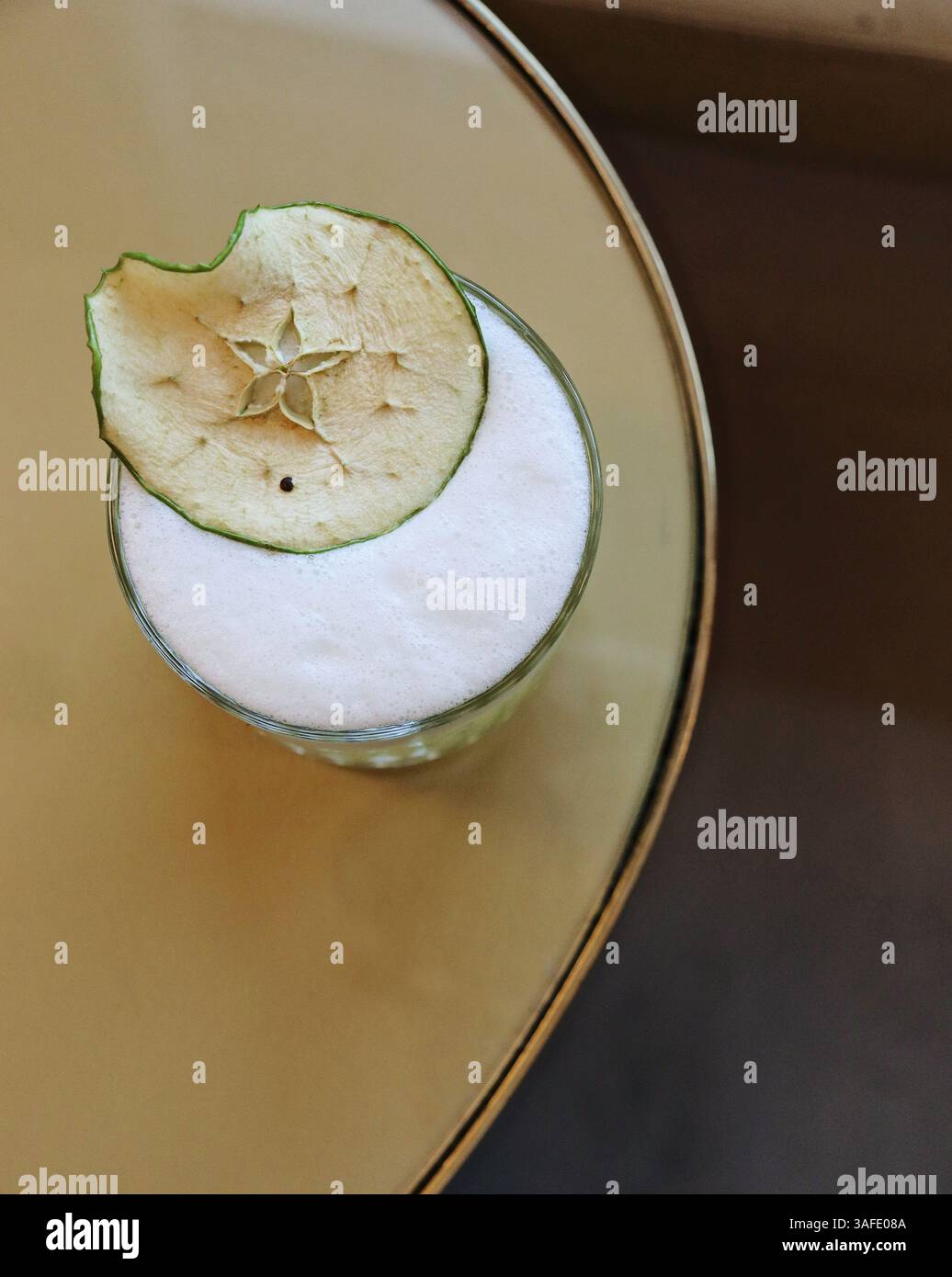Elegante cocktail o mocktail conditi con una delicata schiuma ariosa e guarniti con una fetta di mela secca. Serviti in un bicchiere trasparente su un elegante tavolo dorato, Foto Stock