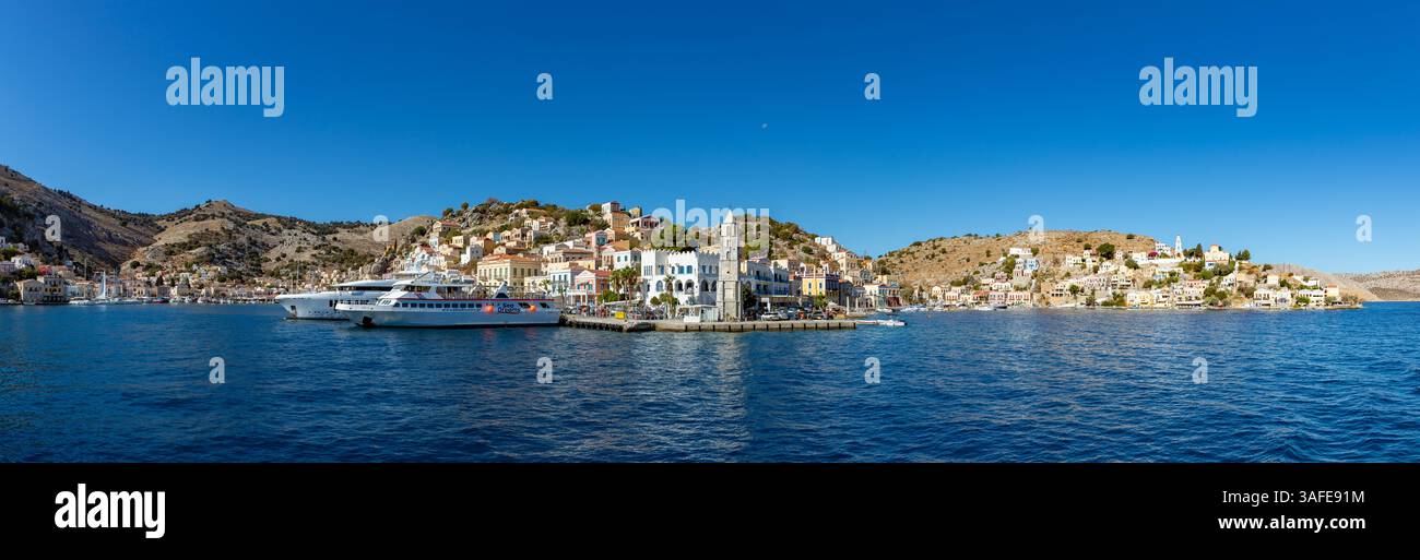 Una foto panoramica della colorata città di Ano Symi sull'isola di Symi, con la torre dell'orologio al centro e le grandi barche sulla sinistra. Foto Stock