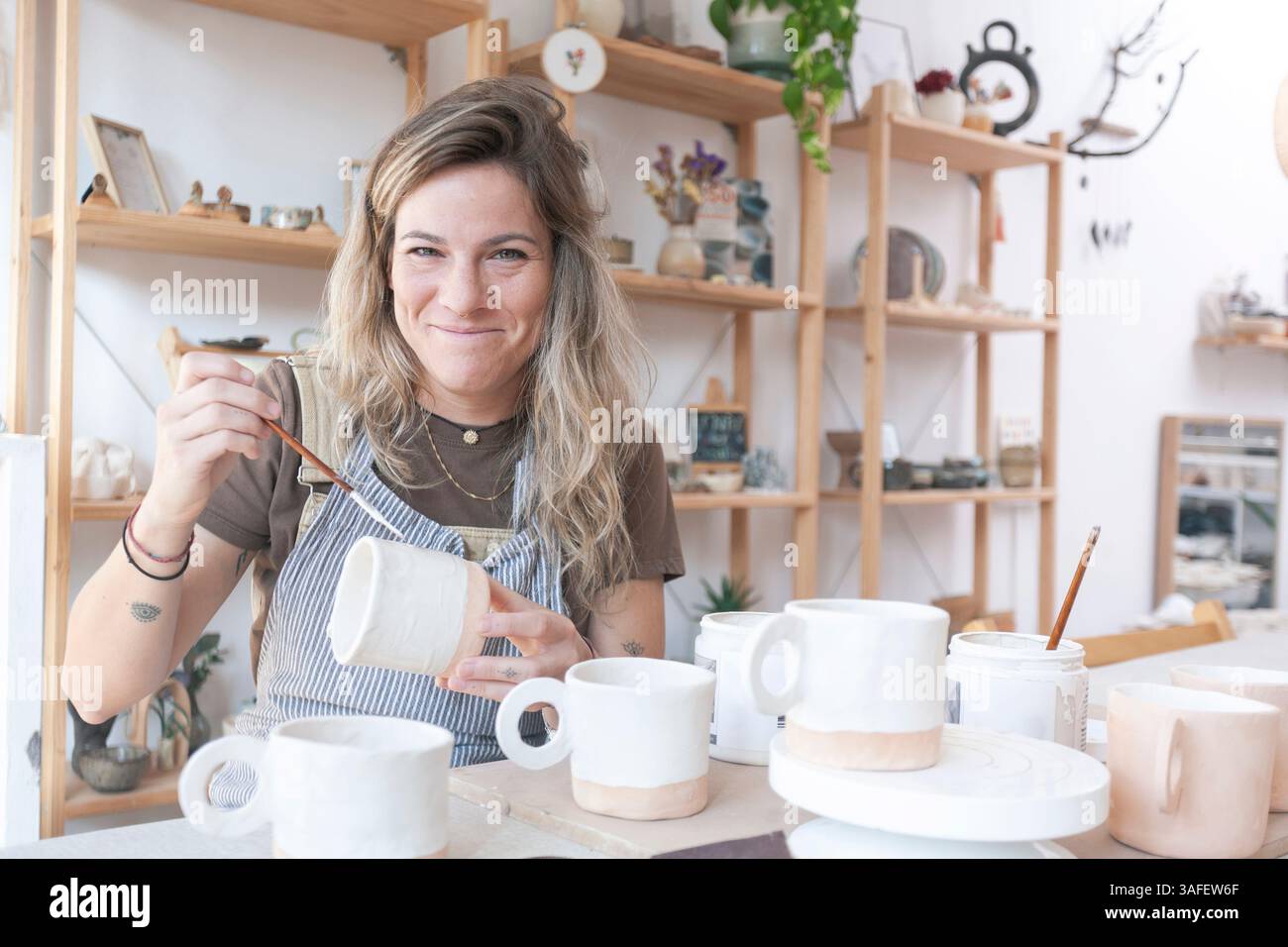 Un'artista di ceramica sorride alla macchina fotografica mentre si mette in pausa dalle sue tazze di argilla, trasudando fiducia e calma nel suo ambiente creativo. Foto Stock