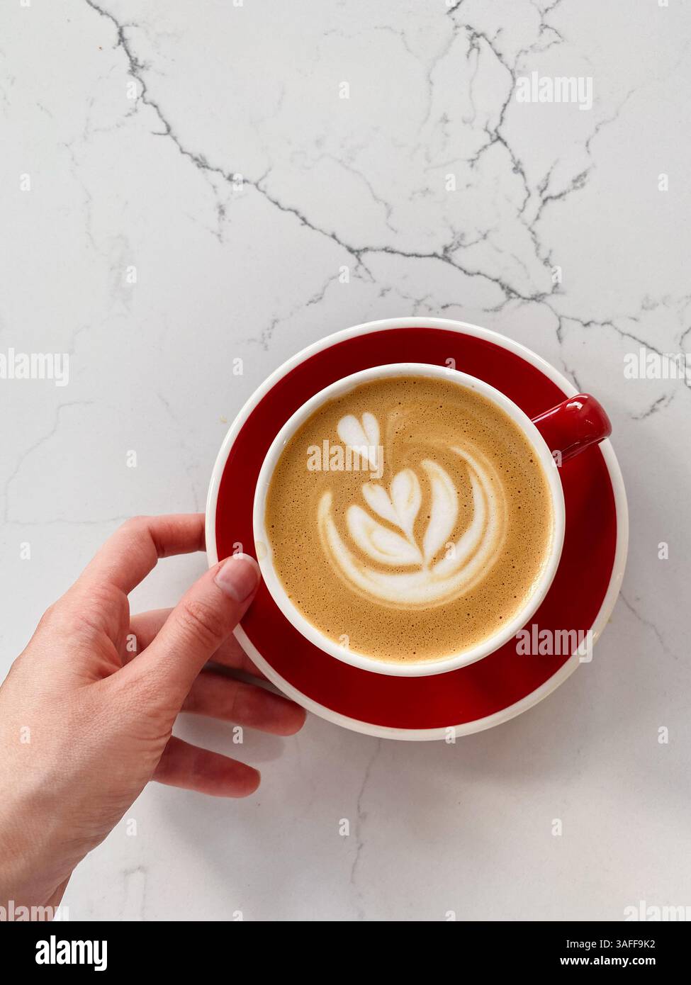 Mano sinistra che regge una tazza di caffè con il cuore contro la superficie del tavolo in marmo bianco Foto Stock