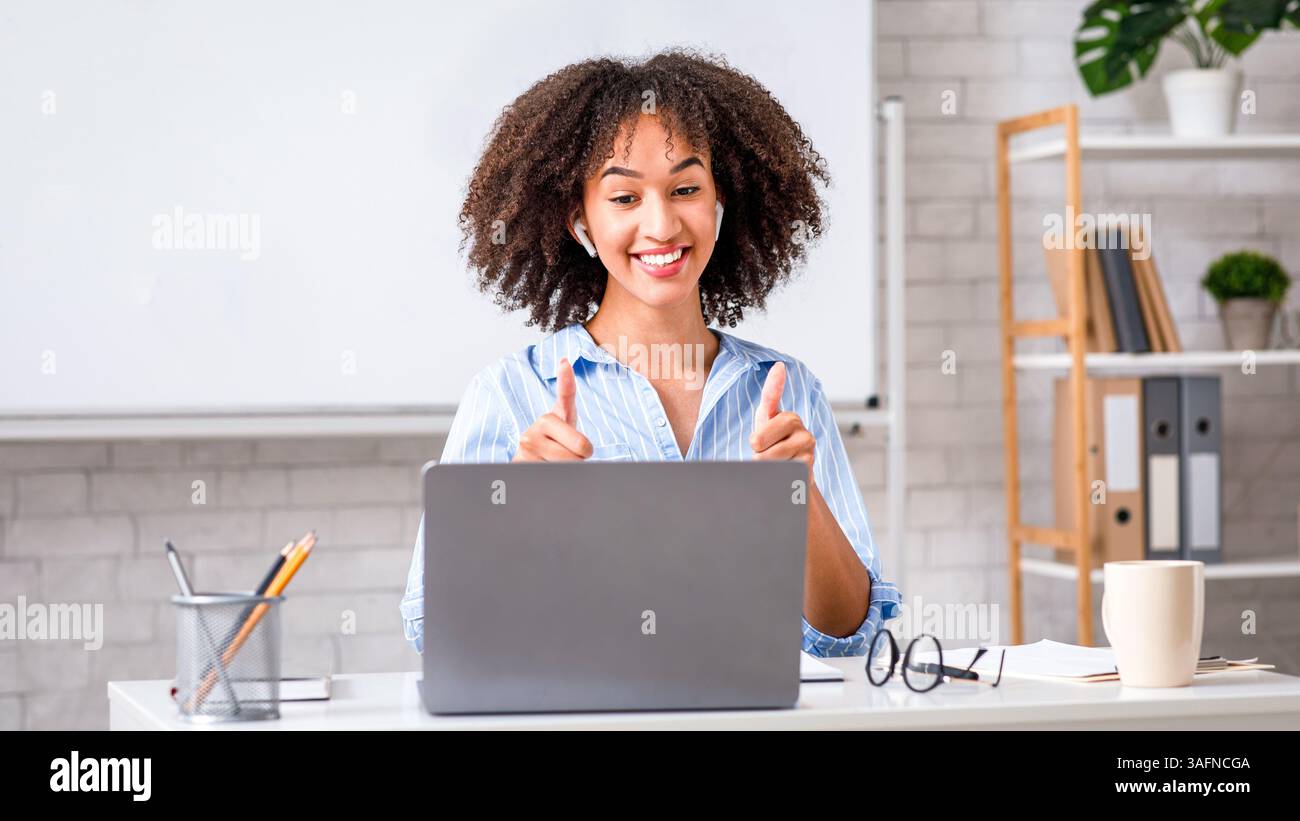 Imparare l'inglese, la protezione della salute e ottimi risultati. Felice giovane afroamericana che mostra i pollici in su e guardando il laptop Foto Stock