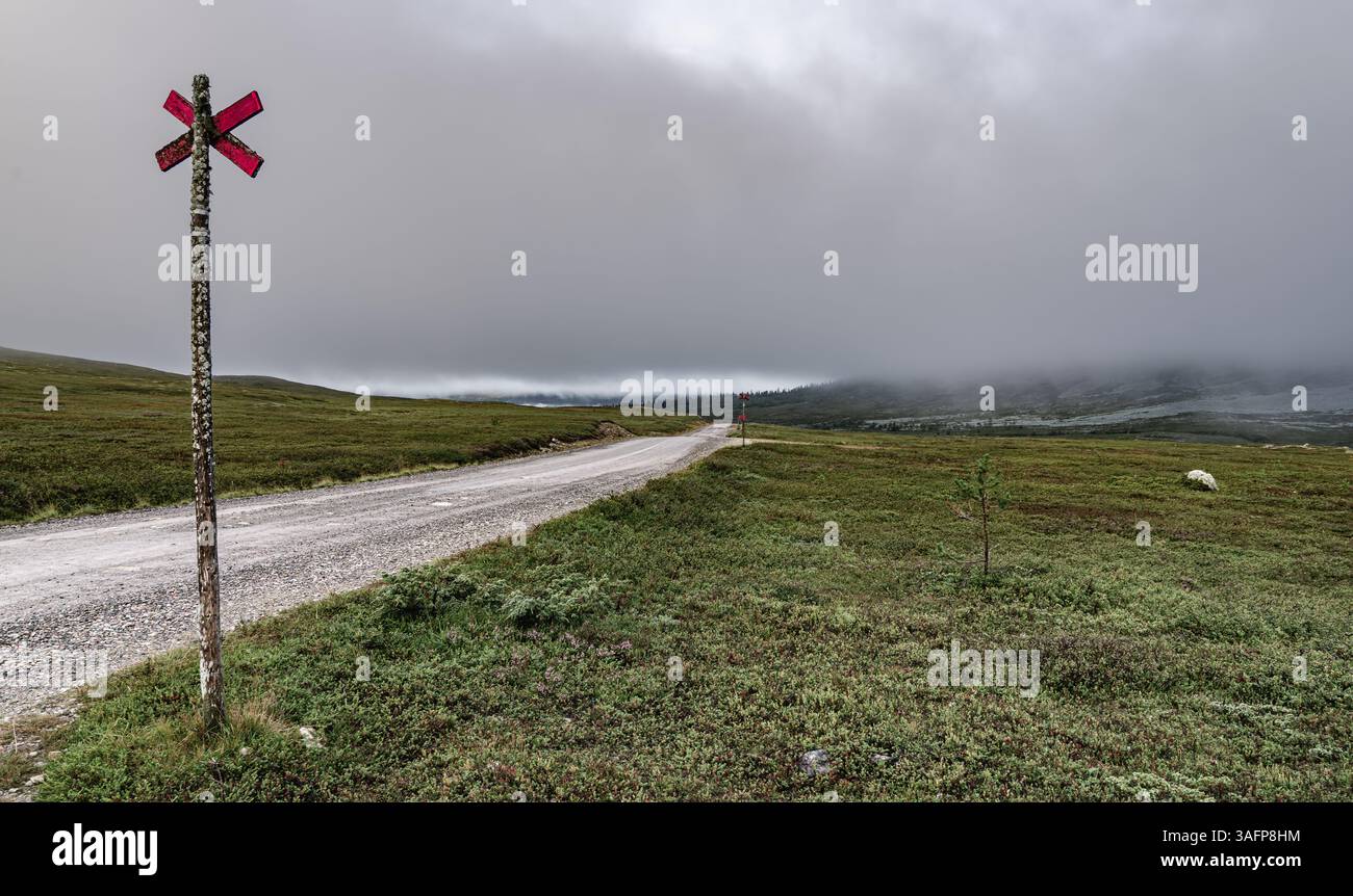 Le nuvole avvolgono le montagne di Idre Dalarna in Svezia, i cartelli rossi delle incroci sui pali si trovano lungo la strada sterrata nella lussureggiante tundra verde Foto Stock