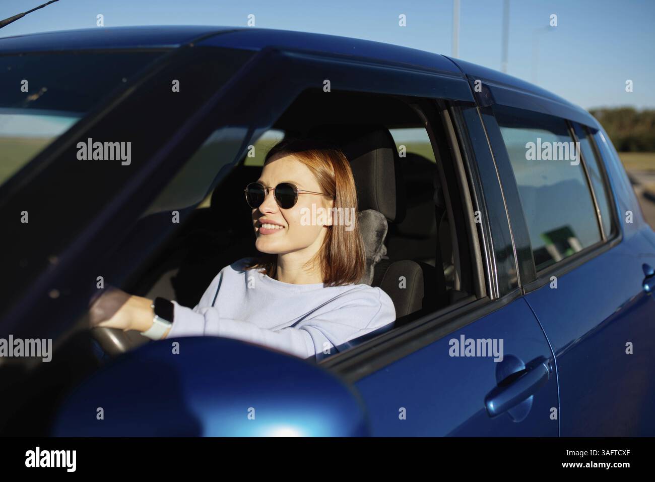 Una donna sorridente che indossa gli occhiali da sole guida un'auto blu sotto il sole Foto Stock