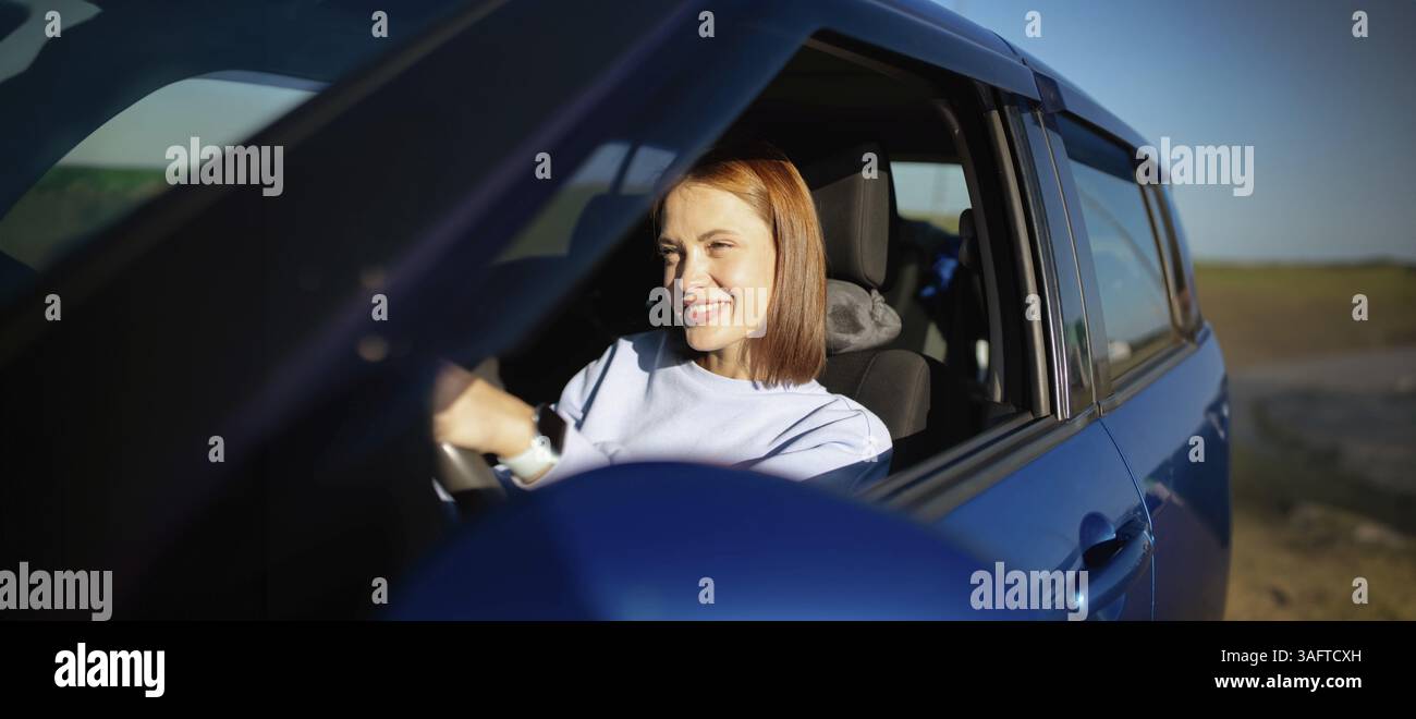 Donna sorridente mentre guida un'auto blu sotto la luce del sole Foto Stock