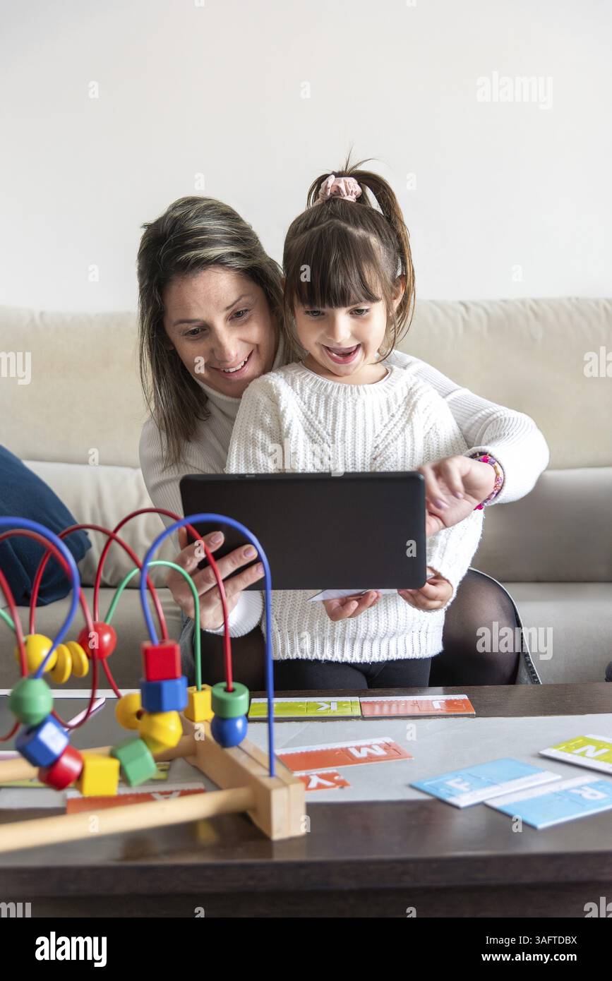 Mamma e figlia si divertono insieme mentre usano un tablet, circondati da giocattoli educativi in un ambiente accogliente Foto Stock