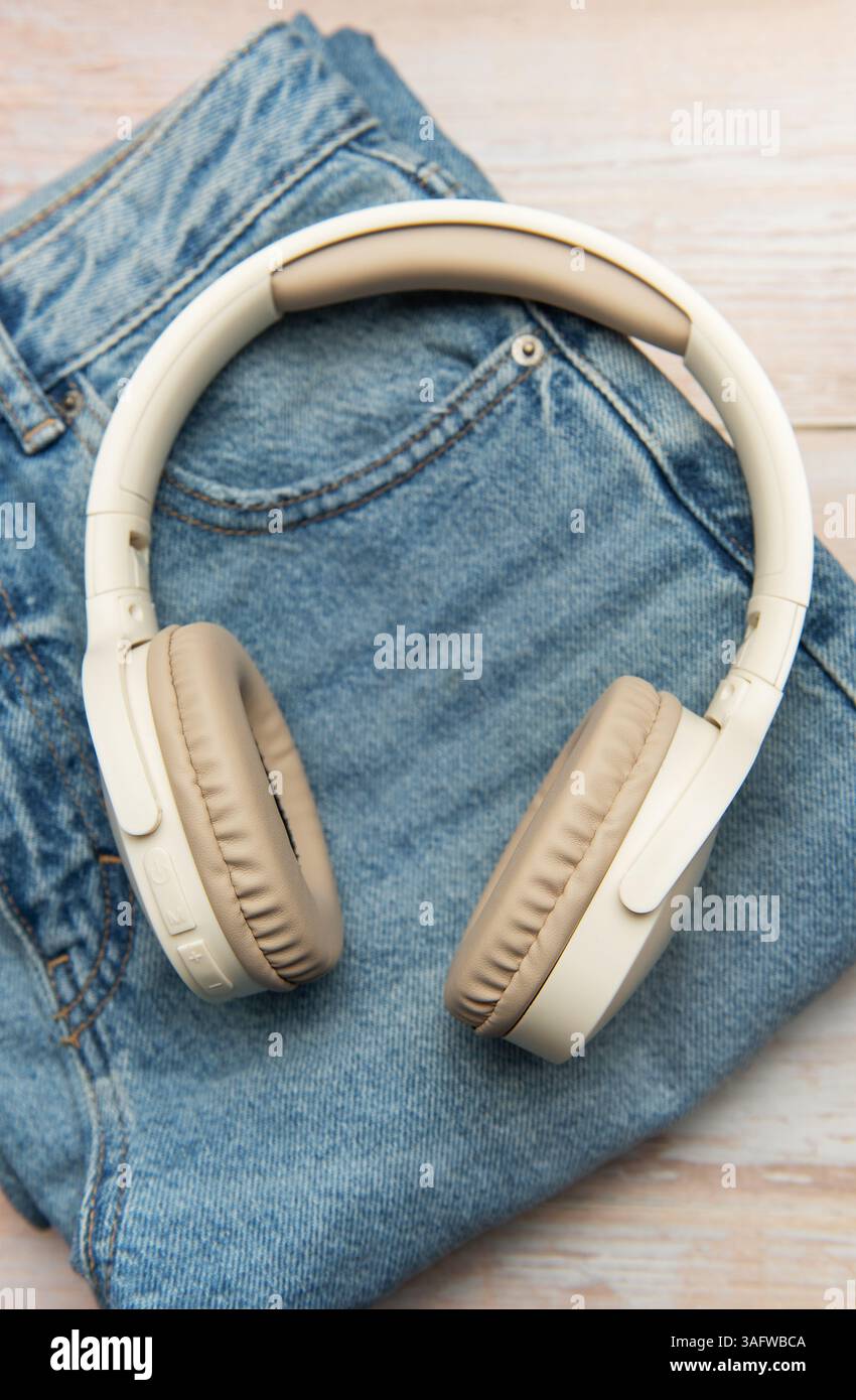 Moderne cuffie wireless posizionate su jeans in denim piegati su uno sfondo di legno chiaro, per creare un'immagine elegante e alla moda Foto Stock