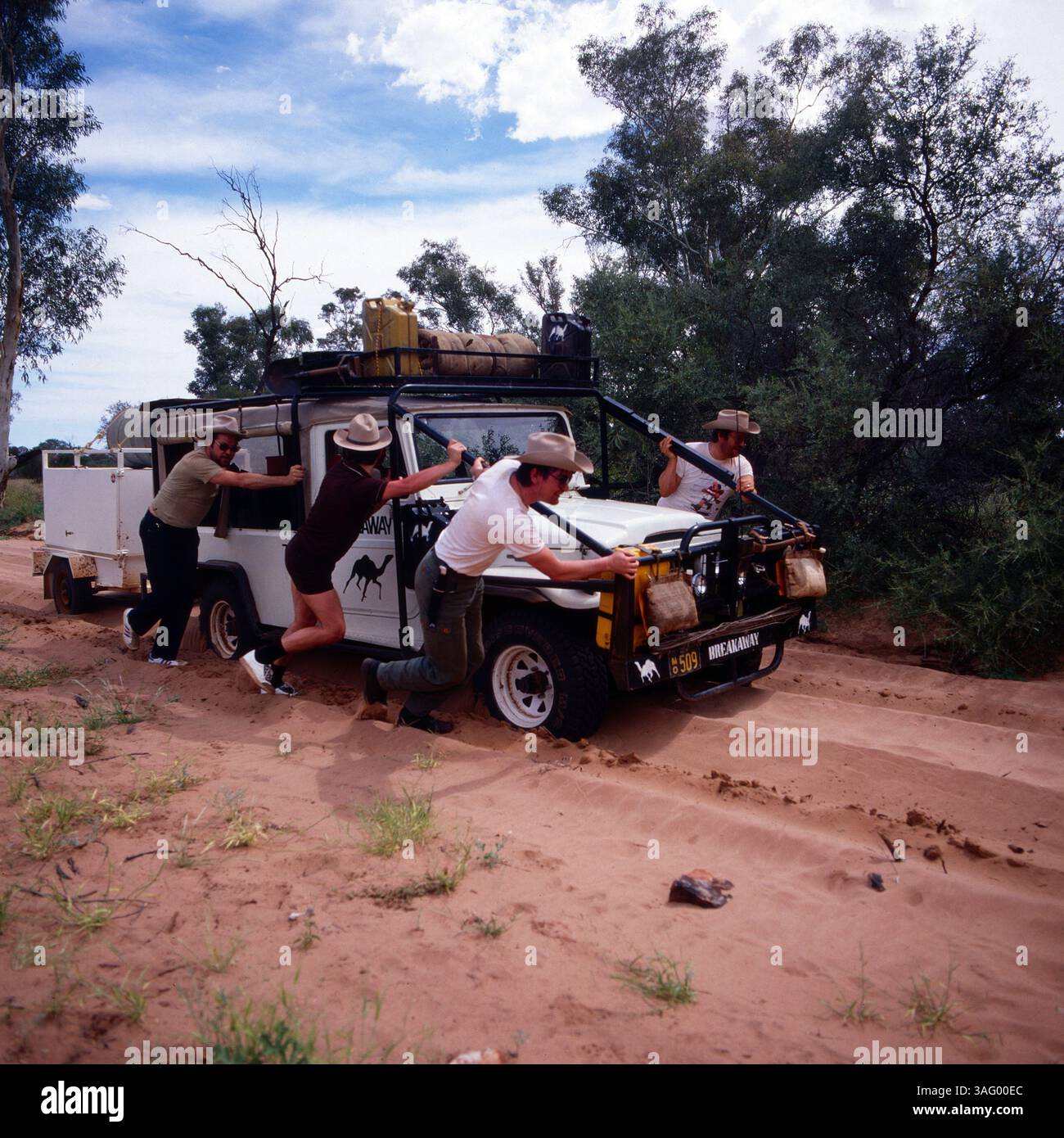 Gruppe von Reisenden oder Abenteurn, Die in der Weite des australischen Outbacks unterwegs sind. IHR Geländewagen ist im tiefen Sand steckengeblieben, und sie kämpfen sich durch, indem sie das Fahrzeug mit vereinten Kräften anschieben. Die Gruppe trägt typische Outback-Kleidung mit Hüten, Shorts und robusten Stiefeln, passend für die heißen und trockenen Bedingungen. DAS Bild vermittelt das Gefühl von Abenteuer, Teamarbeit und Herausforderung in der Wildnis, um 1985. Foto Stock