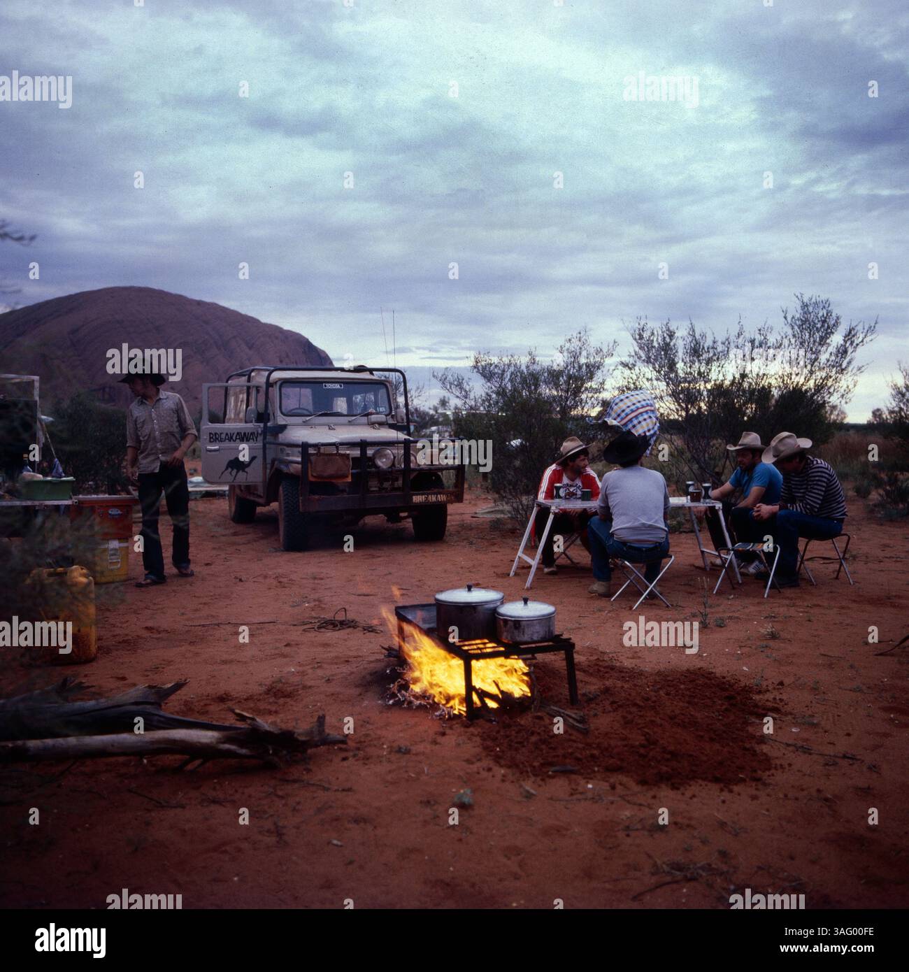 Outback-Campszene in Australien, vermutlich in der Nähe des Uluru (Ayers Rock), der im Hintergrund erkennbar ist. Eine Gruppe von Reisenden Hat ihr Camp mitten in der Wüste aufgeschlagen und sitzt entspannt an einem Klapptisch. Im Vordergrund brennt ein Lagerfeuer, auf dem mehrere Kochtöpfe erhitzt werden. Ein Geländewagen, ausgestattet für Expeditionen, steht bereit, während ein Mann mit Cowboyhut und Outdoor-Kleidung durch das Lager geht. Die Szene vermittelt das Gefühl von Abenteuer, Freiheit und der rauen Schönheit des australischen Outbacks, um 1985. Foto Stock