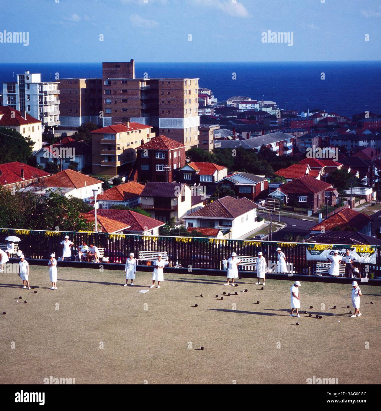 Stadtteil von Sydney, nuovo Galles del Sud, mit einem Grünen Lawn Bowls Club im Vordergrund. Mehrere Ältere Damen in Weißen outfits spielen das traditionelle Spiel Lawn Bowls, während einige Zuschauer auf Bänken sitzen. Im Hintergrund erstreckt sich eine dichte Wohnbebauung mit roten Ziegeldächern und mehrstöckigen Apartmenthäusern, dahinter das tiefblaue Meer. DAS Bild fängt die entspannte Atmosphäre eines sonnigen Nachmittags an der Küste ein, um 1985. Foto Stock