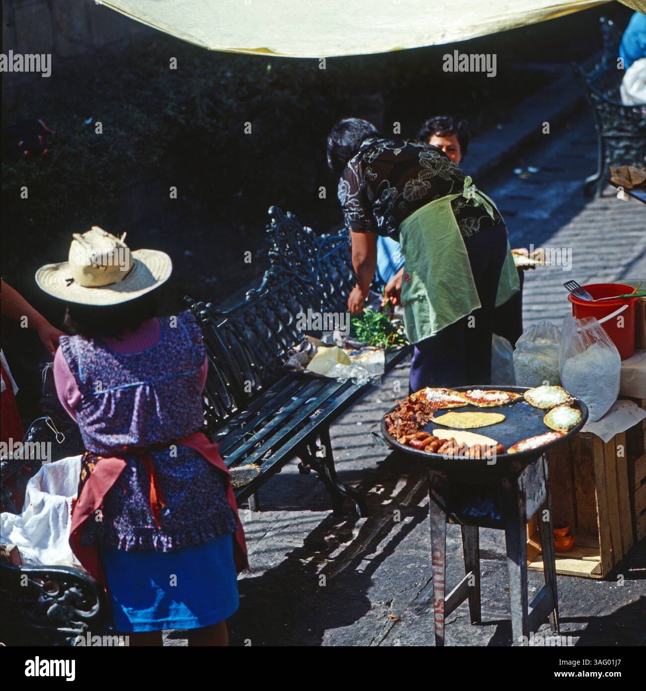 Straßenküche in Mexiko-Stadt: Frauen bereiten an einem improvisierten Stand auf offener Flamme traditionelle mexikanische Speisen zu. Auf einer großen Grillplatte braten tortillas, Fleisch und Gemüse, während frische Zutaten auf einer Bank bereitliegen. Eine Frau mit Strohhut Wartet neben dem Stand, während eine andere in einer Schürze die Zubereitung überwacht. Die Szene zeigt authentisches Straßenessen und mexikanische Esskultur im urbanen Raum, um 1985. Foto Stock