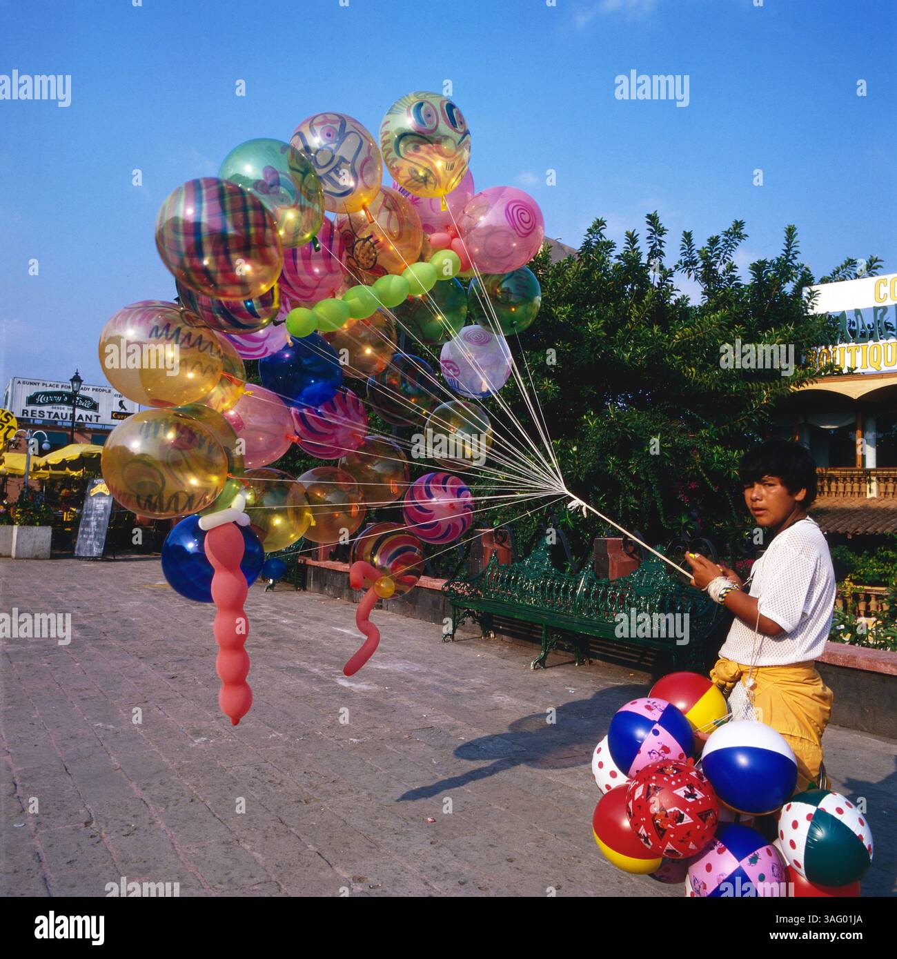 Junger Ballonverkäufer hält eine große Anzahl bunter Luftballons a Mexiko-Stadt. Die Ballons mit verschiedenen Mustern und Farben schweben über ihm, während er sie Fest im Griff Hat. Im Hintergrund sind Grüne Bäume, eine Sitzbank und ein Restaurant mit Sonnenschirmen zu sehen. Die Szene fängt die städtischen eines fröhliche Atmosphäre Platzes ein, um 1985. Foto Stock
