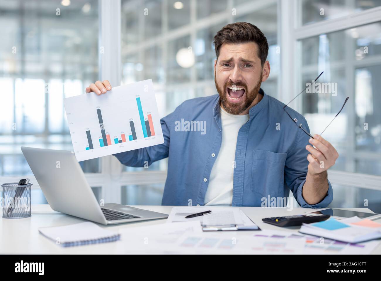 Un uomo d'affari frustrato alla sua scrivania mostra scarsi risultati finanziari su un grafico. Quell'uomo sta tenendo gli occhiali e ha un aspetto disperato. Foto Stock