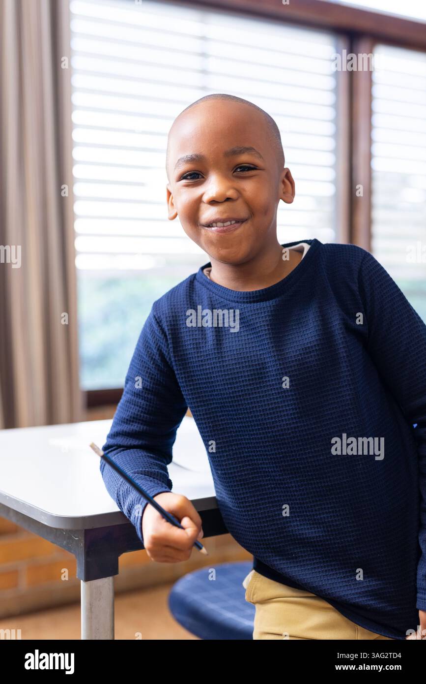 Sorridente ragazzo afroamericano che tiene in mano una matita e si appoggia sulla scrivania in classe scolastica Foto Stock