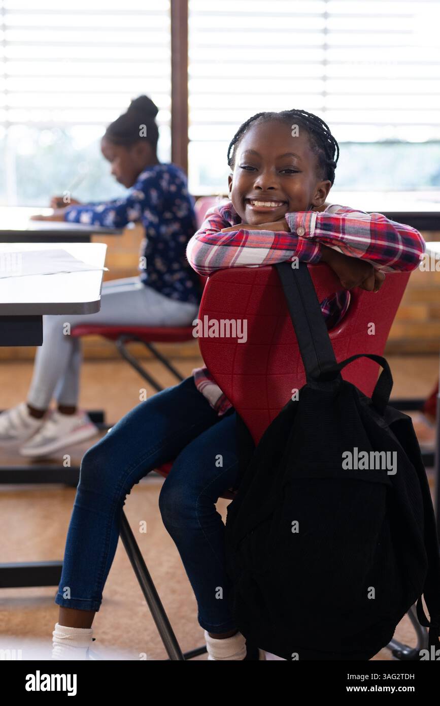 Sorridente ragazza afroamericana seduta su una sedia con lo zaino in classe, godendo una pausa Foto Stock