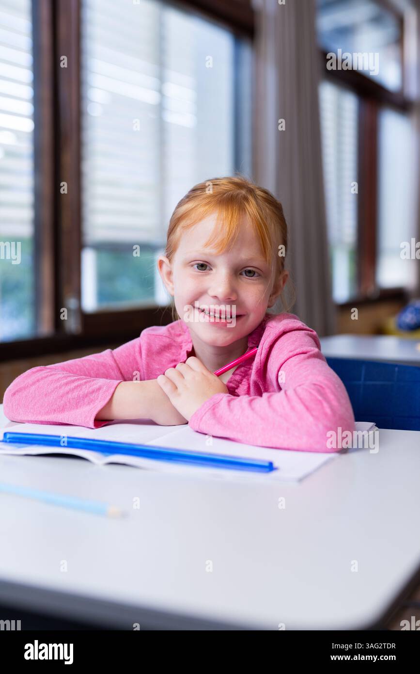 Ragazza sorridente seduta alla scrivania con libro aperto in classe Foto Stock