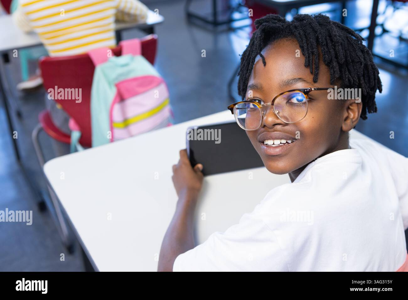 A scuola, ragazzo afroamericano che usa il tablet e sorride mentre seduto alla scrivania Foto Stock
