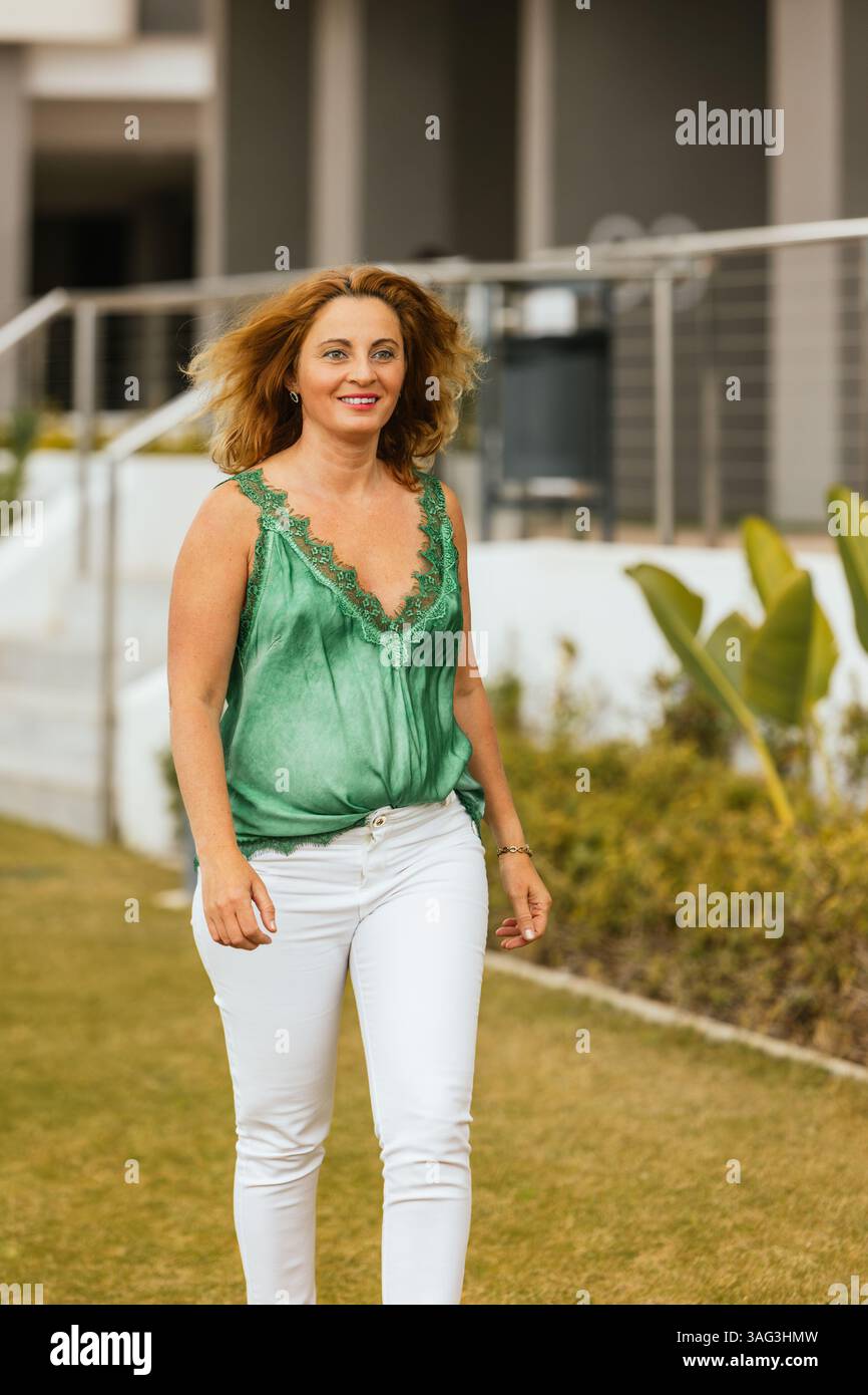 Donna sorridente in un abbigliamento informale che cammina sull'erba fuori da un hotel moderno, godendosi una piacevole giornata in un ambiente di resort tropicale. Vacanze. Stile di vita. Foto Stock