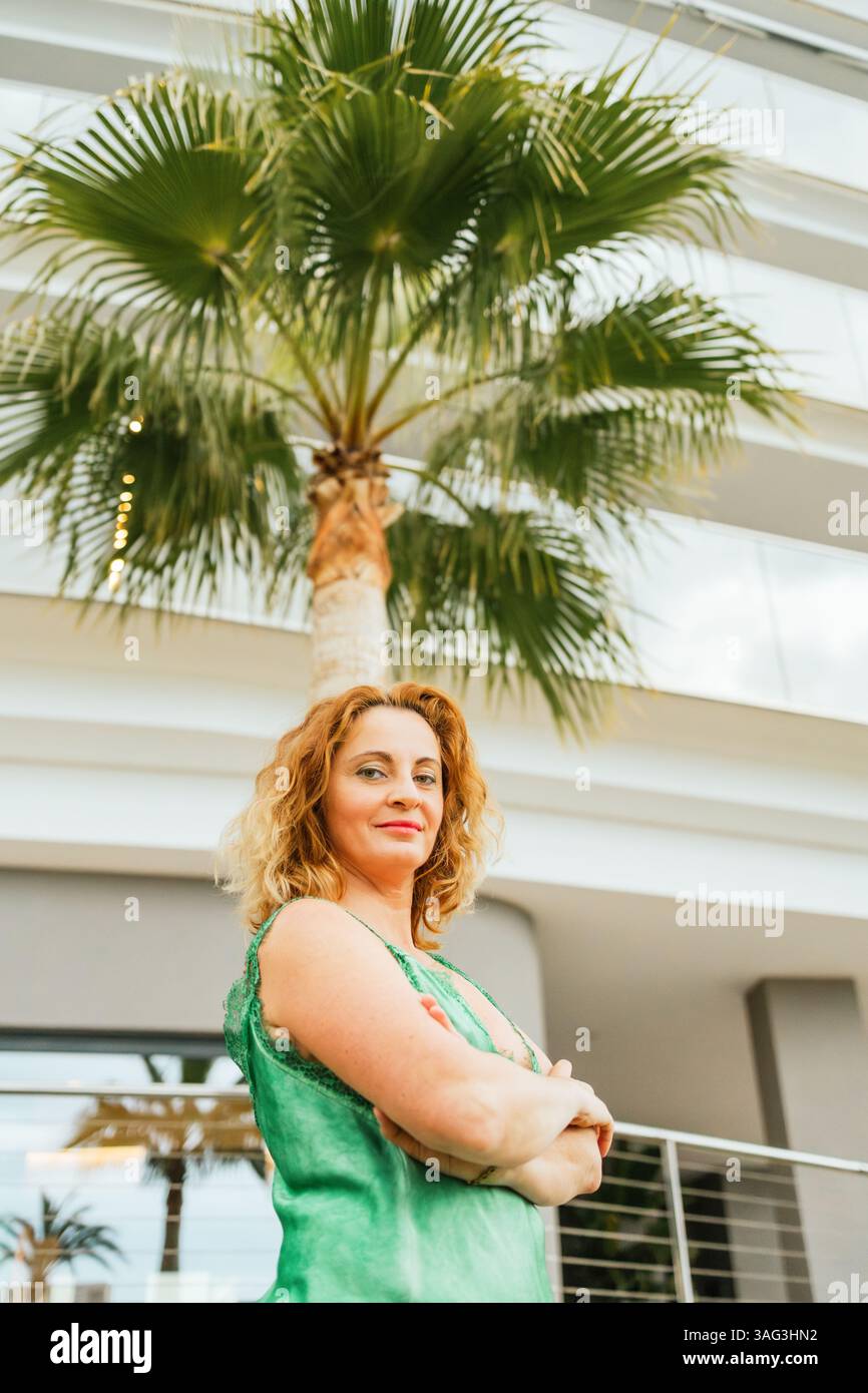 Vista ad angolo basso di una donna sicura di sé con le braccia incrociate, in piedi all'aperto sotto un'alta palma vicino a un moderno edificio alberghiero, trasudando potere e. Foto Stock