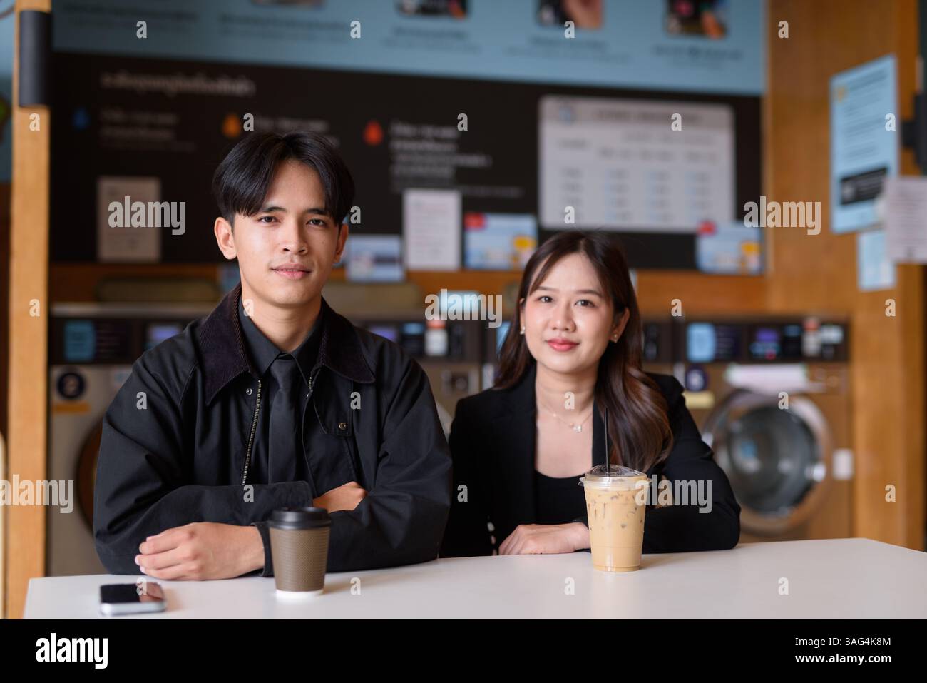Giovane coppia asiatica felice seduta in lavanderia a bere caffè e a usare il telefono Foto Stock