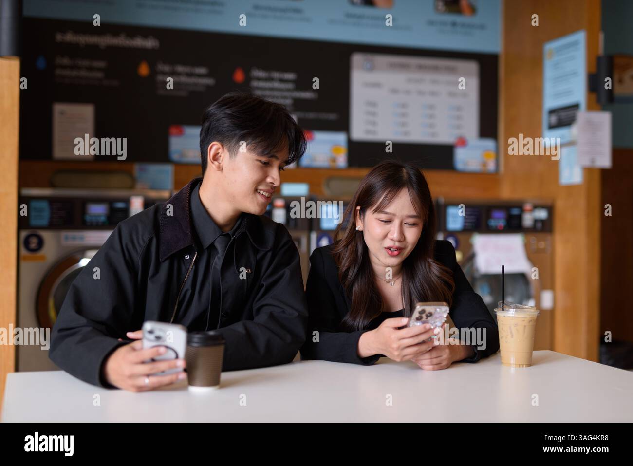Giovane coppia asiatica felice seduta in lavanderia a bere caffè e a usare il telefono Foto Stock