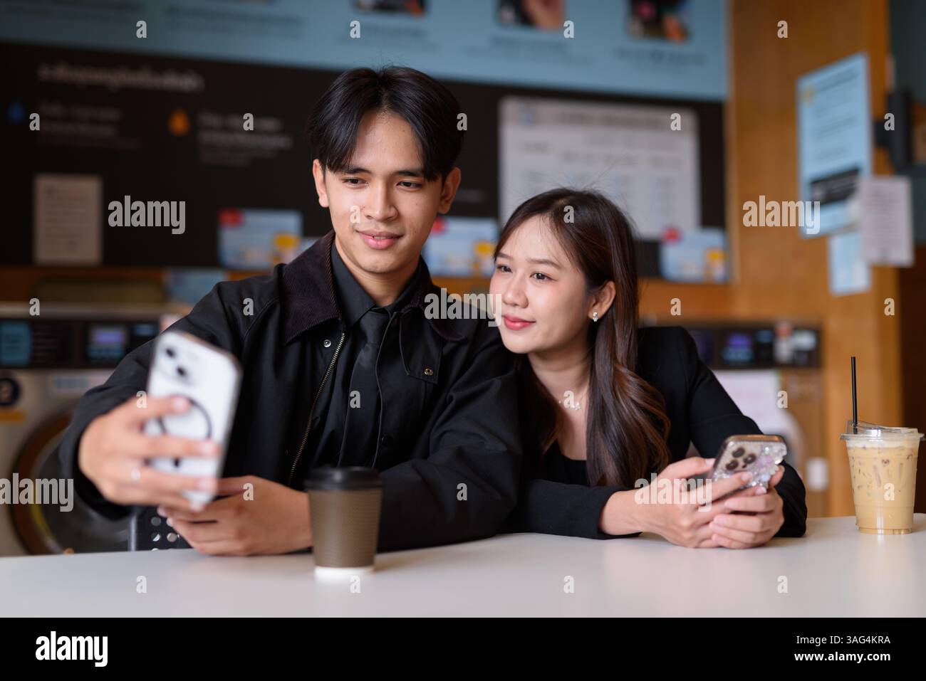 Giovane coppia asiatica felice seduta in lavanderia a bere caffè e a usare il telefono Foto Stock