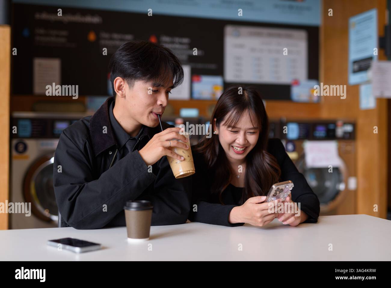 Giovane coppia asiatica felice seduta in lavanderia a bere caffè e a usare il telefono Foto Stock