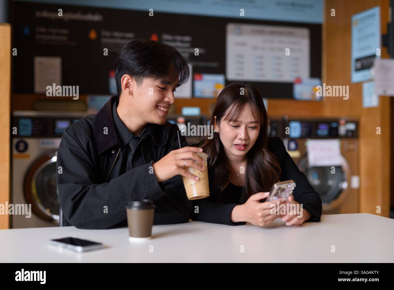 Giovane coppia asiatica felice seduta in lavanderia a bere caffè e a usare il telefono Foto Stock