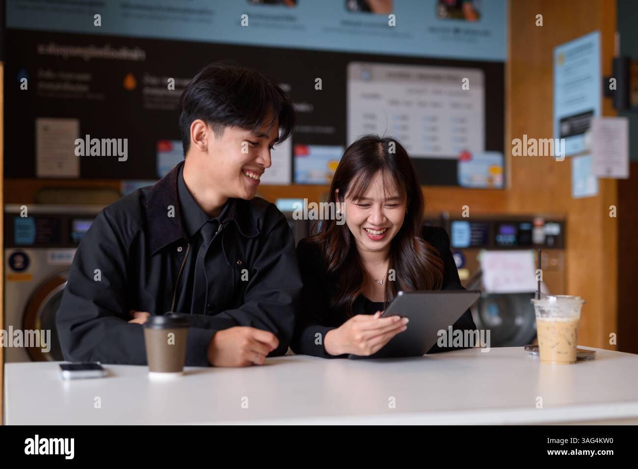 Giovane coppia asiatica felice seduta in lavanderia a bere caffè e a usare un computer tablet Foto Stock